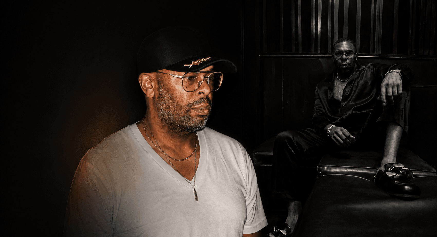 C.L. Smooth in a moody, low-lit portrait — white v-neck, aviators, and a fitted cap.